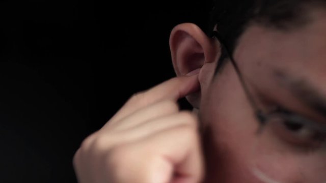 Portrait shot of Caucasian male teenager itching his wax-filled ears isolated on black background.