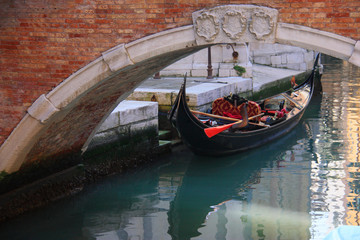 Fototapeta na wymiar Venice, Italy. An empty gondola waiting for passengers or gondolier under a small low bridge. Blue-green colour of water. Day light.