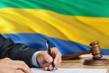 Judge writing on paper in courtroom with Gabon flag background. Wooden gavel of equality theme and...