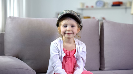 Funny smiling preschooler girl in military cap sitting on sofa, little patriot