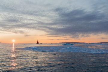 A small boat among icebergs. Sailboat cruising among floating icebergs in Disko Bay glacier during...