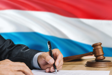 Judge writing on paper in courtroom with Luxembourg flag background. Wooden gavel of equality theme...