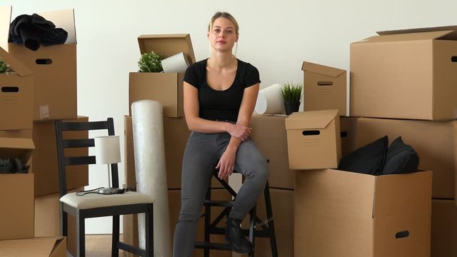 A moving woman sits on a chair and looks seriously at the camera in an empty apartment, surrounded by cardboard boxes