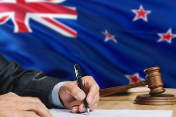 Judge writing on paper in courtroom with New Zealand flag background. Wooden gavel of equality...