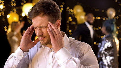 Young man suffering migraine at party massaging temples, annoying music, health