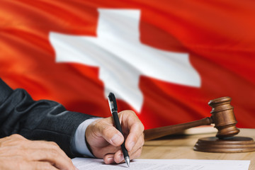 Judge writing on paper in courtroom with Switzerland flag background. Wooden gavel of equality...