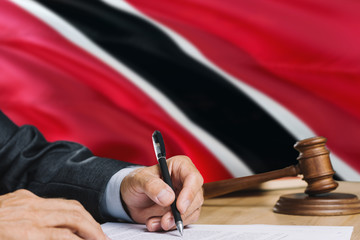 Judge writing on paper in courtroom with Trinidad And Tobago flag background. Wooden gavel of equality theme and legal concept.