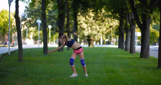 Beautiful Caucasian girl of seven years in sporty clothes practicing wheel gymnastic exercise on one hand on the grass in a city park on a sunny summer day. Slow motion