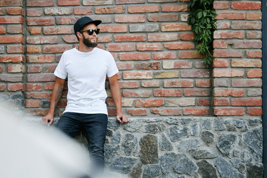 Hipster handsome male model with beard wearing white blank t-shirt and a baseball cap with space for your logo or design in casual urban style