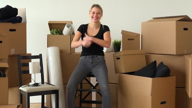 A happy moving woman sits on a chair in an empty apartment, dances and smiles at the camera, surrounded by cardboard boxes