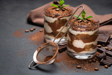 Classic tiramisu dessert in a glass cup, strainer and pieces of chocolate on dark concrete background