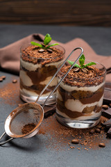 Classic tiramisu dessert in a glass cup, strainer and pieces of chocolate on dark concrete background