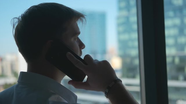 Young promising businessman talking on the phone against the background of the panoramic windows of his office. The view from the window on the city and high-rise buildings.