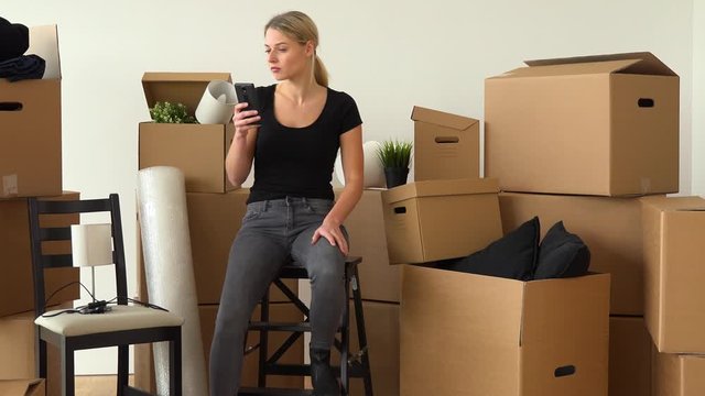A moving woman sits on a chair in an empty apartment and takes pictures of cardboard boxes surrounding her