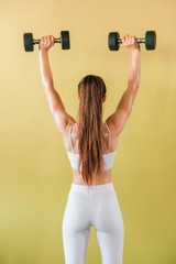 Athletic bodybuilder woman with dumbbells. Beautiful brunette girl with muscles lifting weights on yellow background.