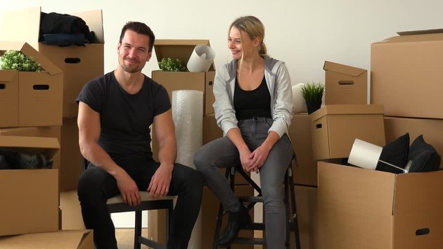 A happy moving couple sits on chairs in an empty apartment and nods at the camera with a smile, surrounded by cardboard boxes