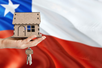 Woman hand holding wooden house and keys with hand. Chile flag with concept of rent, purchase, insurance, building real estate, eco house.