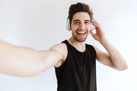 Happy young strong sports man posing isolated over white wall background listening music with headphones take a selfie by camera.