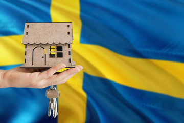 Woman hand holding wooden house and keys with hand. Sweden flag with concept of rent, purchase, insurance, building real estate, eco house.