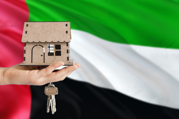 Woman hand holding wooden house and keys with hand. United Arab Emirates flag with concept of rent,...