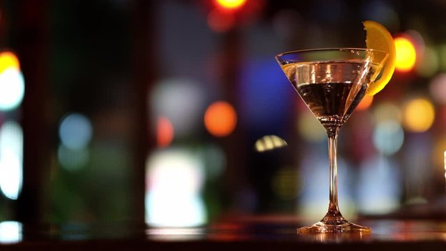 Close up a glass of martini cocktail with orange slice on table in the bar. Defocused colorful lights background