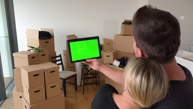 SAME A moving couple looks at a tablet with green screen in an empty apartment, a pile of cardboard boxes in the background