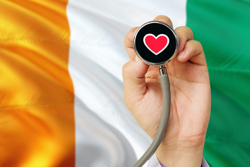 Doctor holding stethoscope with red love heart. National Cote D'Ivoire flag background. Healthcare system concept, medical theme.