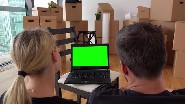 A moving couple looks at a notebook with green screen in an empty apartment, a pile of cardboard boxes in the background