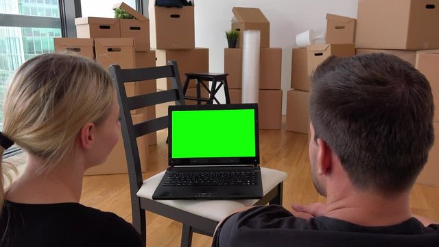 A moving couple looks at a notebook with green screen and talks in an empty apartment, a pile of cardboard boxes in the background
