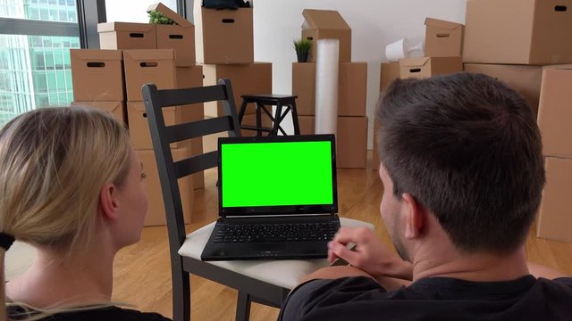 A moving couple looks at a notebook with green screen and talks in an empty apartment, he occasionally works on it, a pile of cardboard boxes in the background