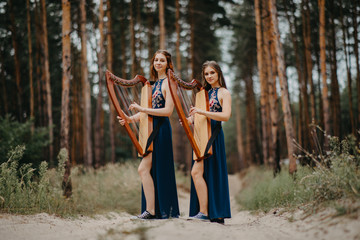 Two women harpists stand at forest and play harps against a background of pines.
