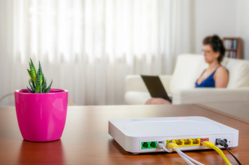 Modem router on a table in a living room. A woman using a laptop while sitting on the sofa is in background. - obrazy, fototapety, plakaty