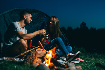 young couple having fun while camping at night outdoor. grilling sausages and drinking beer