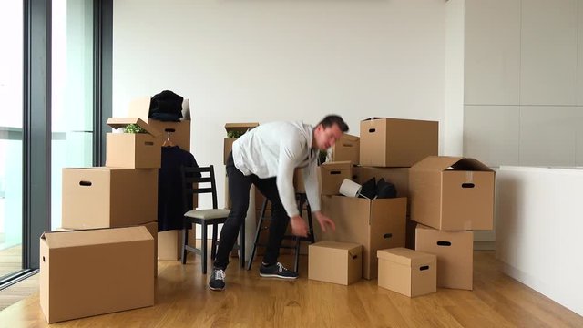 A moving man catches cardboard boxes thrown by someone off the camera and puts them to the pile of more boxes in an empty apartment