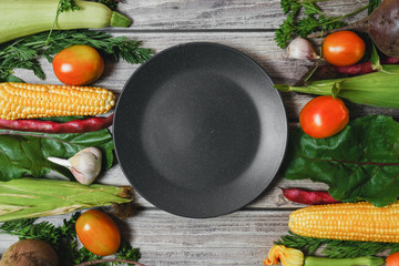 Creative layout made of corn, tomatoes, zucchini, carrot, beetroot, garlic, bean and black plate on wooden background. Food concept. Flat lay, top view. Free space for your text.