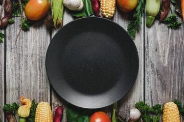 Assorted fresh vegetables on wooden background. Corn, tomatoes, zucchini, carrot, beetroot, garlic, bean and black plate. Food concept. Flat lay, top view, copy space