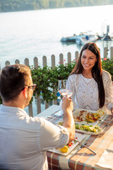 Romantic young couple making a toast, celebrating their anniversary or birthday in a restaurant by water. Husband and wife eating fish dinner in outdoor restaurant by the river
