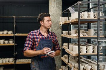 Middle aged male potter in apron notes something in his notebook while counts pottery on shelves.