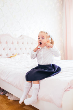 Little adorable blonde girl in school uniform and white golfs siting on a ber in her bedroom and shockes about first day in the school.