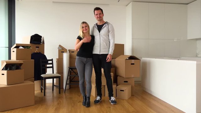 A friendly moving couple stands in an empty apartment and beckons to the camera in a gesture of invitation - cardboard boxes in the background
