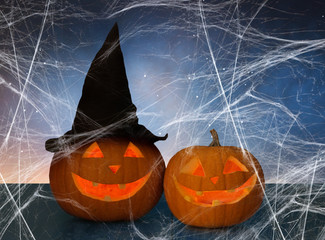 holidays, halloween and decoration concept - two pumpkins or jack o lanterns in witch hat on table and spiderweb over starry night sky background