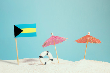 Miniature flag of Bahamas on beach with colorful umbrellas and life preserver. Travel concept, summer theme.