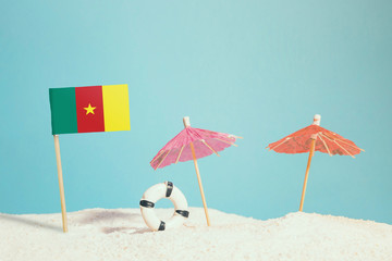 Fototapeta premium Miniature flag of Cameroon on beach with colorful umbrellas and life preserver. Travel concept, summer theme.