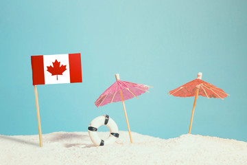 Miniature flag of Canada on beach with colorful umbrellas and life preserver. Travel concept, summer theme.