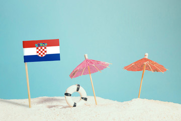 Miniature flag of Croatia on beach with colorful umbrellas and life preserver. Travel concept, summer theme.