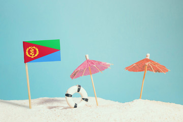 Miniature flag of Eritrea on beach with colorful umbrellas and life preserver. Travel concept, summer theme.