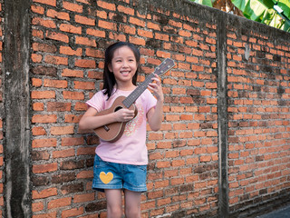 Little asian child play the ukulele, old brick wall background