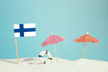 Miniature flag of Finland on beach with colorful umbrellas and life preserver. Travel concept, summer theme.