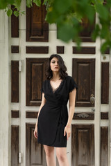 Beautiful brunette girl, wearing a black dress with a deep neckline, poses while standing on the street at an old wooden door. Advertising, fashion, commercial design. Copy space