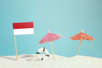 Miniature flag of Indonesia on beach with colorful umbrellas and life preserver. Travel concept, summer theme.